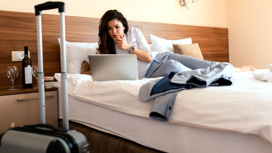 woman on hotel bed with laptop and cabin suitcase in foreground